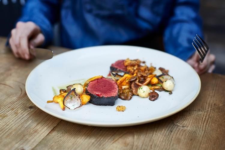 Person sat behind aplate of meat and vegetables, holding knife and fork Person sat behind aplate of meat and vegetables, holding knife and fork