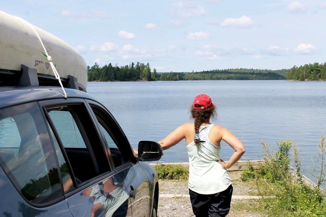 woman leans against a car with a canoe strapped to the top as she looks out onto Temagami lake woman leans against a car with a canoe strapped to the top as she looks out onto Temagami lake