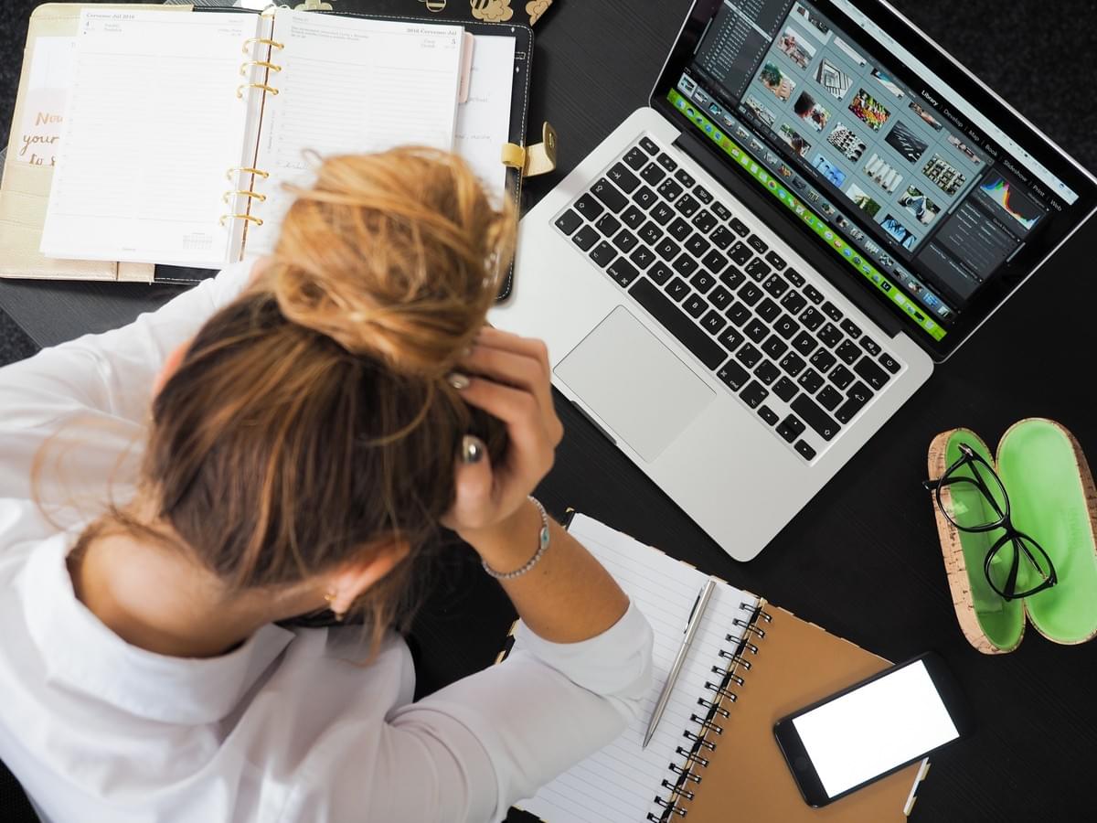 woman sitting at her work desk in front of laptop and books with her head in her hands visibly stressed woman sitting at her work desk in front of laptop and books with her head in her hands visibly stressed