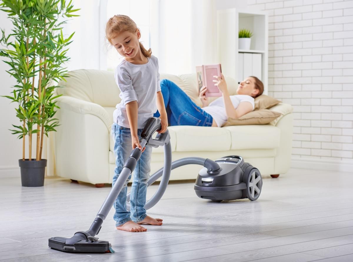 Petite fille passant l'aspirateur dans la maison pendant que sa maman lit un livre, allongée sur le canapé en arrière plan, lâcher prise, tâche ménagère, enfant Petite fille passant l'aspirateur dans la maison pendant que sa maman lit un livre, allongée sur le canapé en arrière plan, lâcher prise, tâche ménagère, enfant