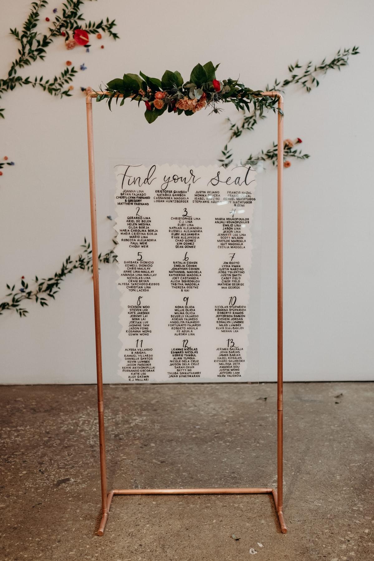 Seating chart on clear acrylic sign. Lettering is done in black ink on a white background, and the sign hangs on a copper frame. Seating chart on clear acrylic sign. Lettering is done in black ink on a white background, and the sign hangs on a copper frame.