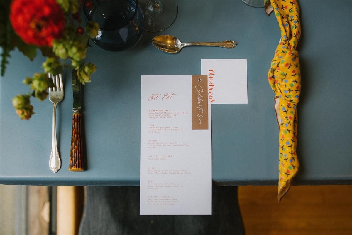 Bird's eye view of a table arrangement featuring a printed menu and hand-lettered placecard from calligrapher Swiftly Scripted. Bird's eye view of a table arrangement featuring a printed menu and hand-lettered placecard from calligrapher Swiftly Scripted.