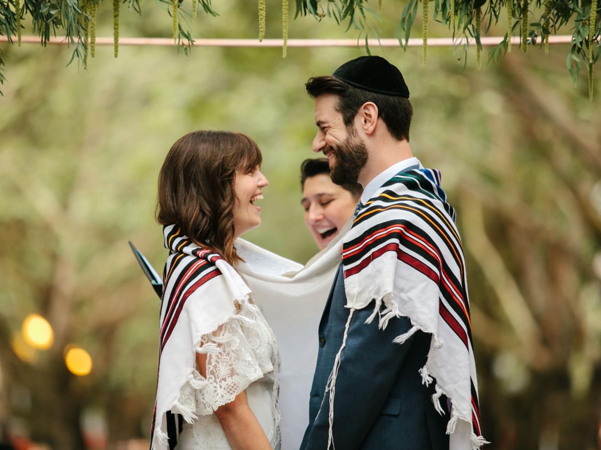 Couple draped in a tallit shares a laugh while the rabbi speaks during their wedding ceremony. Couple draped in a tallit shares a laugh while the rabbi speaks during their wedding ceremony.