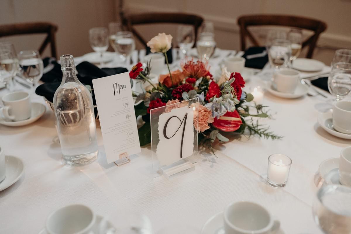 Tropical floral centerpiece in red, peach, and blue, complemented by a table number, menu, and linens in crisp black and white. Tropical floral centerpiece in red, peach, and blue, complemented by a table number, menu, and linens in crisp black and white.
