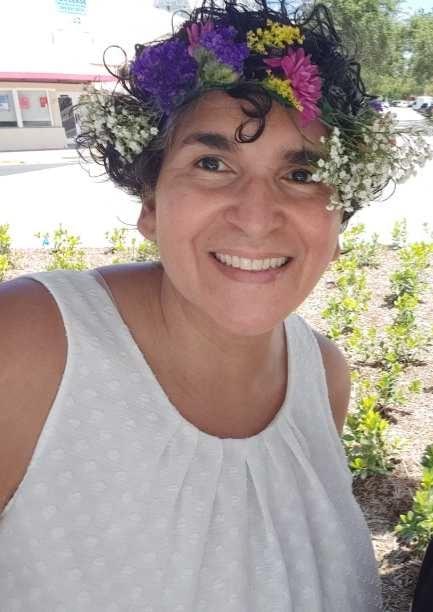 The author. A smiling very tan woman with short curly hair. She is wearing a crown of flowers - Baby's breath on either side with purple, yellow and fuschia flowers in the center. She is wearing a white tank shirt with embroided white flowers. She is surrounded by shurbs and greenry in the background. The author. A smiling very tan woman with short curly hair. She is wearing a crown of flowers - Baby's breath on either side with purple, yellow and fuschia flowers in the center. She is wearing a white tank shirt with embroided white flowers. She is surrounded by shurbs and greenry in the background.