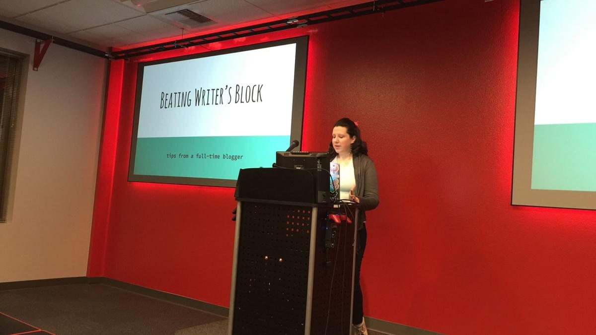 Kitty presenting in front of a bright red wall with a slide that says Beating Writer's Block Kitty presenting in front of a bright red wall with a slide that says Beating Writer's Block