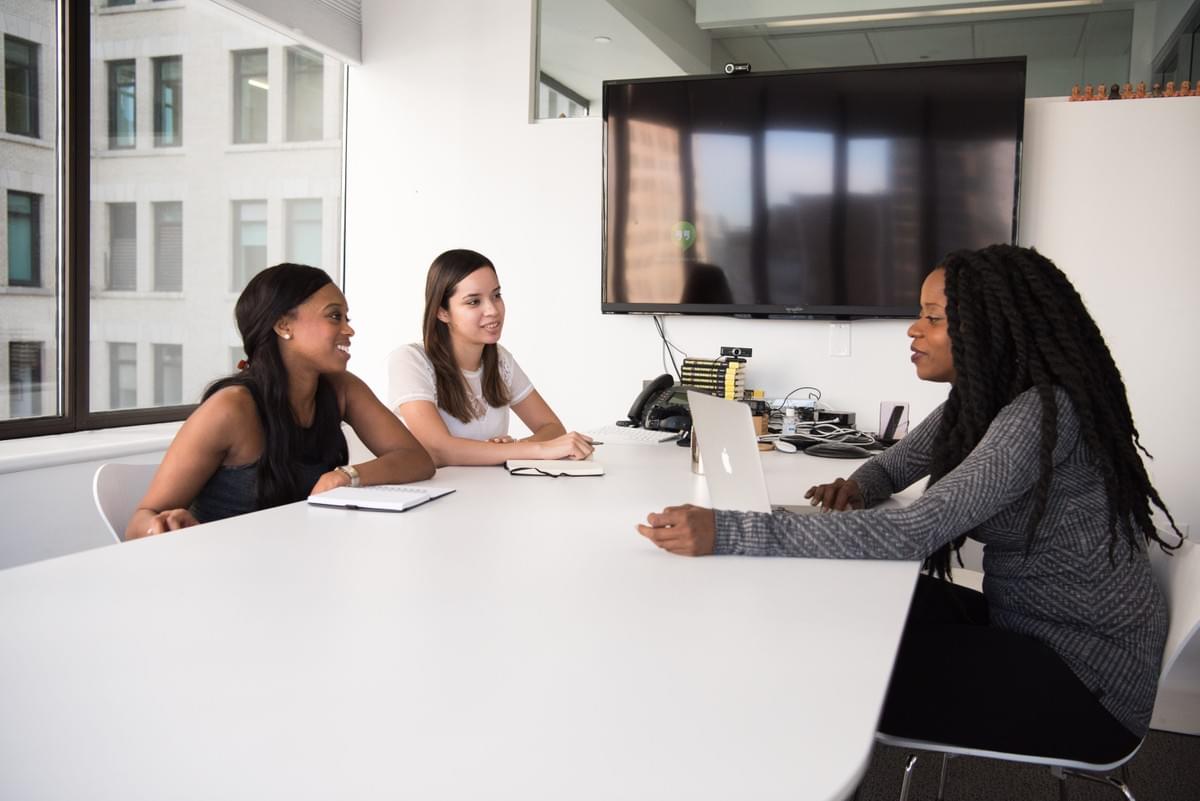 Women, including women of color, sitting at a conference room table with notebooks and a Macbook, apparently collaborating at work Women, including women of color, sitting at a conference room table with notebooks and a Macbook, apparently collaborating at work