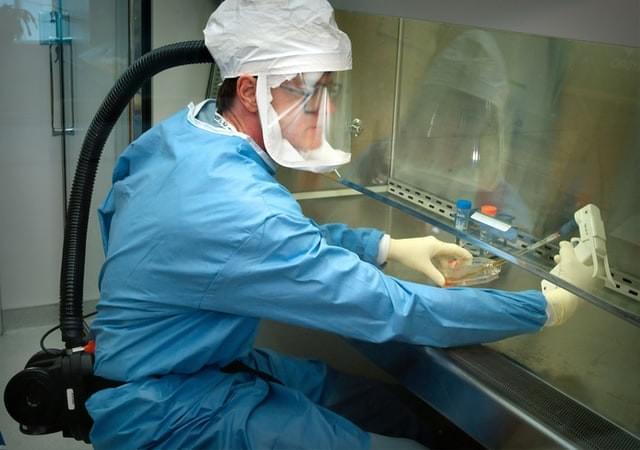 An image of a medical researcher in a blue suit with gloves, a hood, a ventilator, and clear face mask working with a pipette An image of a medical researcher in a blue suit with gloves, a hood, a ventilator, and clear face mask working with a pipette