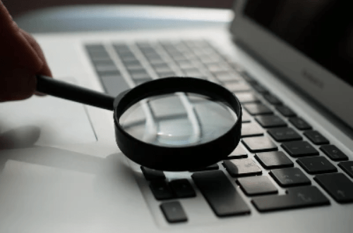 A closeup of a laptop. There is a hand holding a magnifying glass closely to the keyboard. A closeup of a laptop. There is a hand holding a magnifying glass closely to the keyboard.