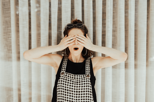 A woman is standing in front of a soft cream background. She has her mouth open in surprise and is covering her eyes with her hands. A woman is standing in front of a soft cream background. She has her mouth open in surprise and is covering her eyes with her hands.