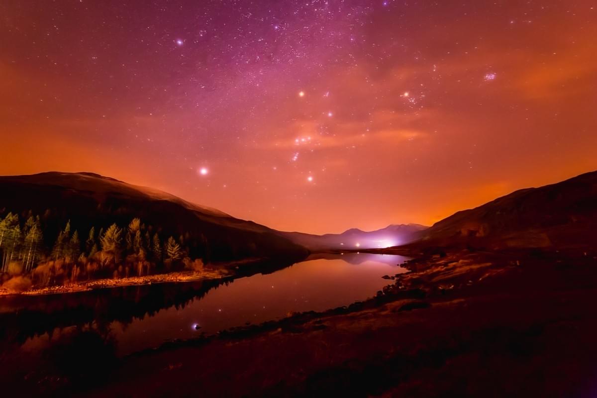 Image of a lake with hills on either side. The starry sky, with the constellation of Orion, is blushed pink, and that pink glow touches the hills and the calm waters of the lake. Here is the profound Oneness of our Universe, and the spiritual peace of our Planet Earth. Image of a lake with hills on either side. The starry sky, with the constellation of Orion, is blushed pink, and that pink glow touches the hills and the calm waters of the lake. Here is the profound Oneness of our Universe, and the spiritual peace of our Planet Earth.