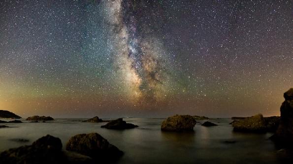 A seashore at night. The sea is calm and is studded with rocks. The sky is filled with stars, and the Milky Way is resplendent. A seashore at night. The sea is calm and is studded with rocks. The sky is filled with stars, and the Milky Way is resplendent.