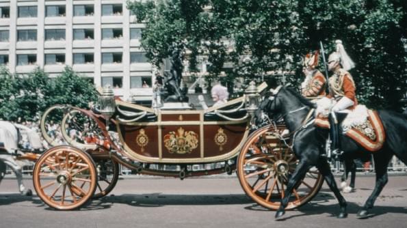 A royal carriage accompanied by mounted Lifeguards passes along a London Street. The carriage is open, and we see the former Queen Mother smiling and waving. A royal carriage accompanied by mounted Lifeguards passes along a London Street. The carriage is open, and we see the former Queen Mother smiling and waving.