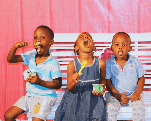 Three adorable children sit on a white bench in front of a pink, wooden building. They are all eating ice cream. Three adorable children sit on a white bench in front of a pink, wooden building. They are all eating ice cream.