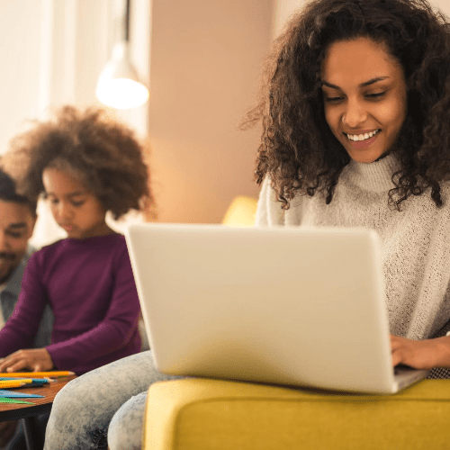 A mom works on her couch with her laptop open as her husband plays with their child. A mom works on her couch with her laptop open as her husband plays with their child.