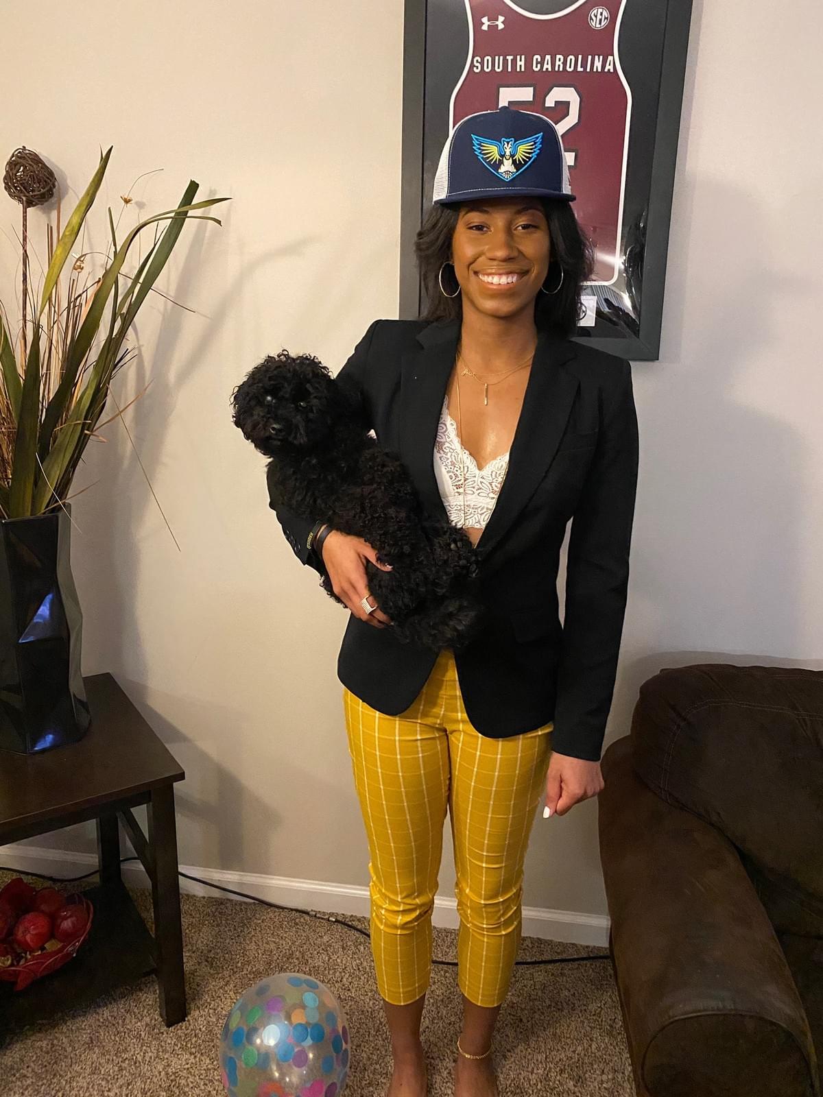 Photo of Ty on draft night wearing Wings hat, black blazer over white lace bralette and yellow checkered pants. She holds a small fluffy black dog (Gucci) in one arm. Photo of Ty on draft night wearing Wings hat, black blazer over white lace bralette and yellow checkered pants. She holds a small fluffy black dog (Gucci) in one arm.