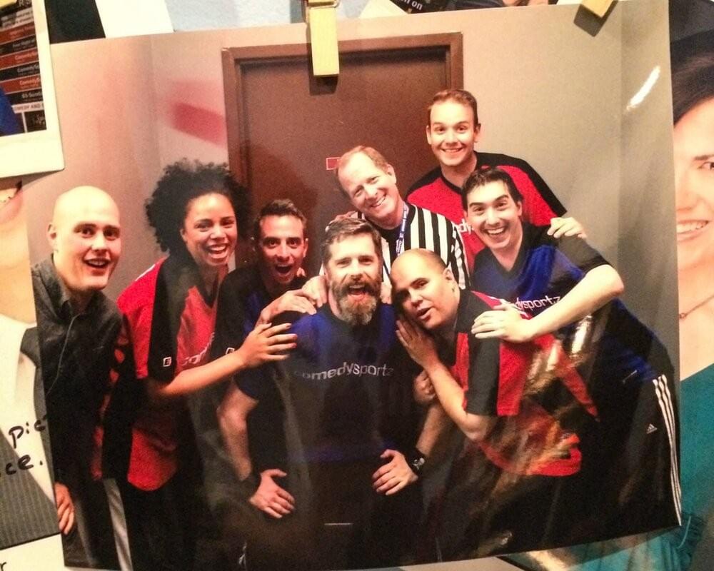 Steven (back right) backstage after a ComedySportz Chicago improv ‘match’ as part of their professional ensemble. Image description: An image of a photo pegged to a pin board. A group of improvisors in sports-style tshirts, smiling at the camera. Steven is in the back (he is the tallest). Steven (back right) backstage after a ComedySportz Chicago improv ‘match’ as part of their professional ensemble. Image description: An image of a photo pegged to a pin board. A group of improvisors in sports-style tshirts, smiling at the camera. Steven is in the back (he is the tallest).