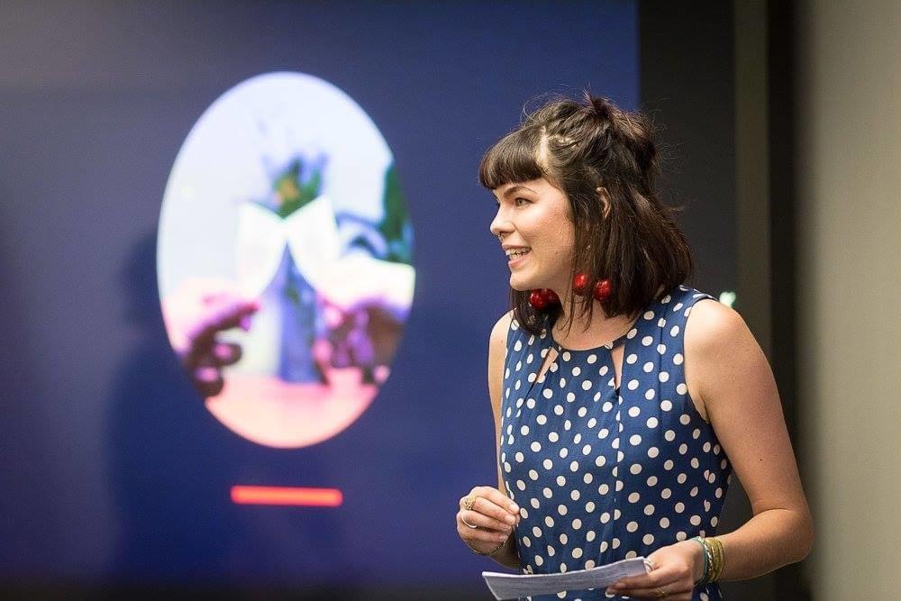 Young Pākehā woman with shoulder length dark hair and a blue and white polka dot top presents in front of a blury background slide. Young Pākehā woman with shoulder length dark hair and a blue and white polka dot top presents in front of a blury background slide.