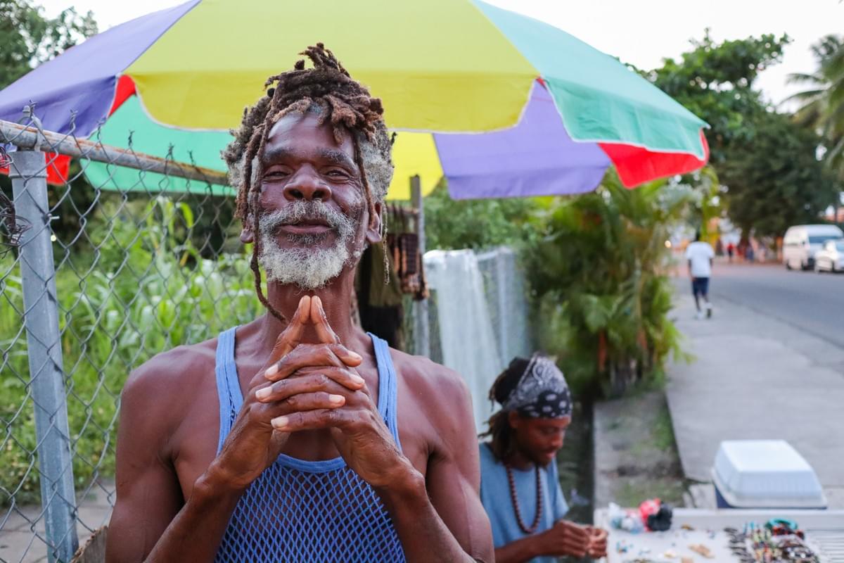 Rasta elder smiling in Saint Lucia while a man sits at a table and makes jewelry Rasta elder smiling in Saint Lucia while a man sits at a table and makes jewelry