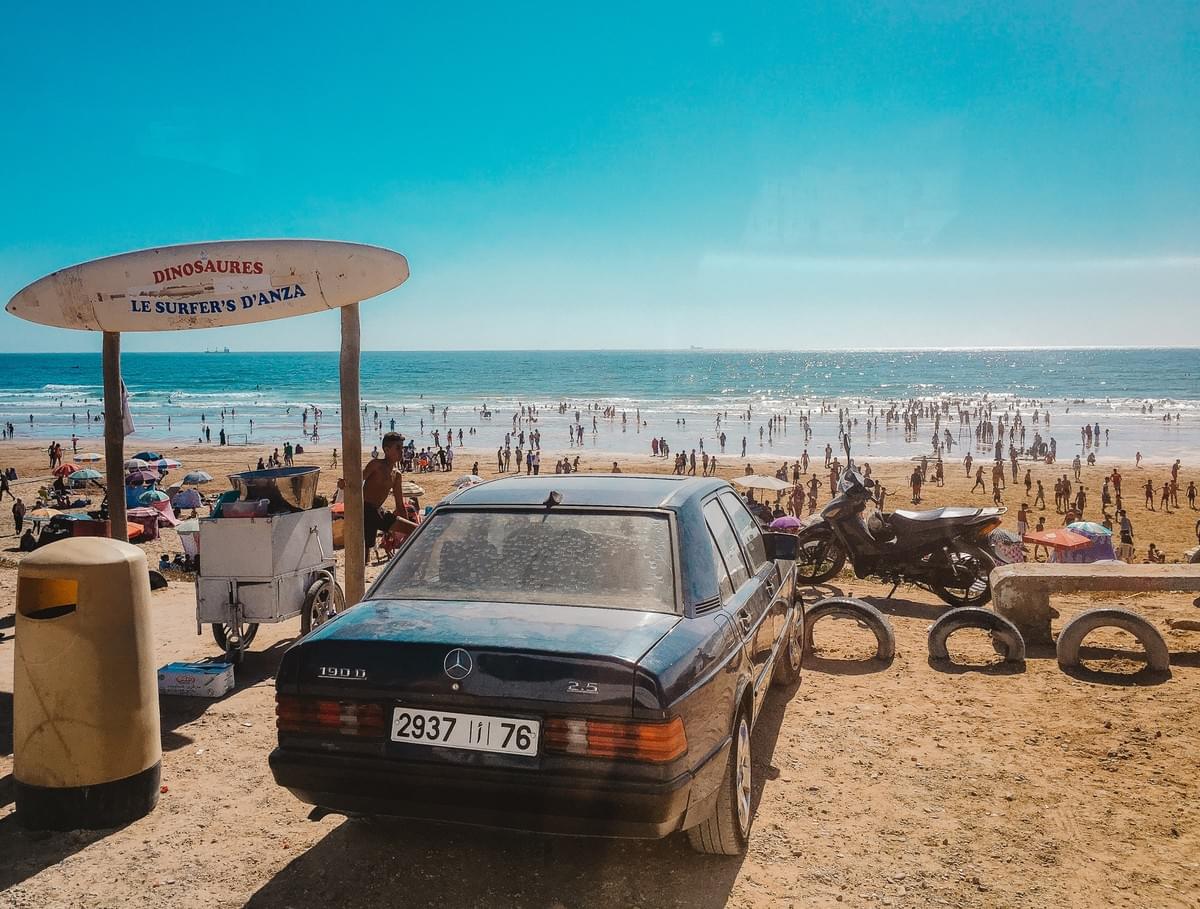 Vintage mercedes, motorcycle and tires parked at the beach next to a trashcan and display surfboard that reads dinosaures le surfer's d'anza with people swimming in the ocean at taghazout bay, agadir, morocco Vintage mercedes, motorcycle and tires parked at the beach next to a trashcan and display surfboard that reads dinosaures le surfer's d'anza with people swimming in the ocean at taghazout bay, agadir, morocco