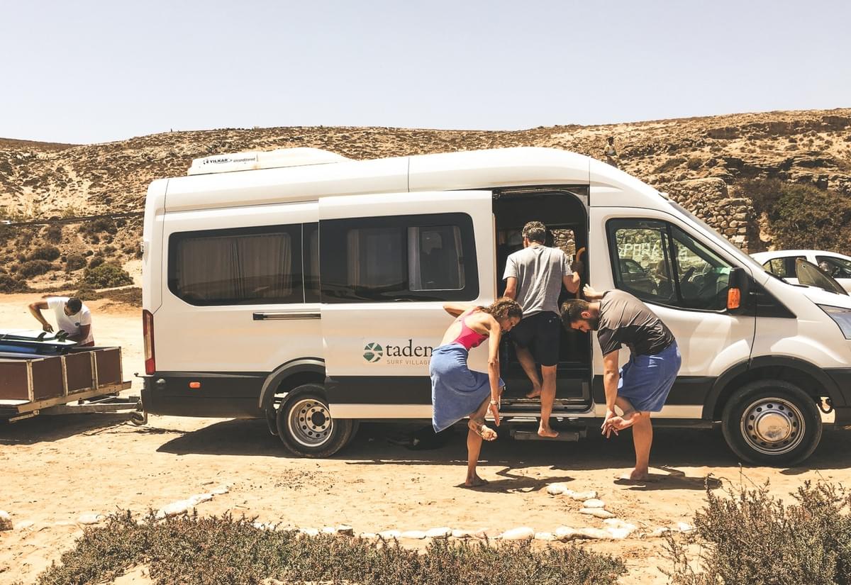 A man unloading surfboards in the back of a truck and people dusting sand of their feet infant of a tadenga shuttle van parked in Tamri, morocco A man unloading surfboards in the back of a truck and people dusting sand of their feet infant of a tadenga shuttle van parked in Tamri, morocco