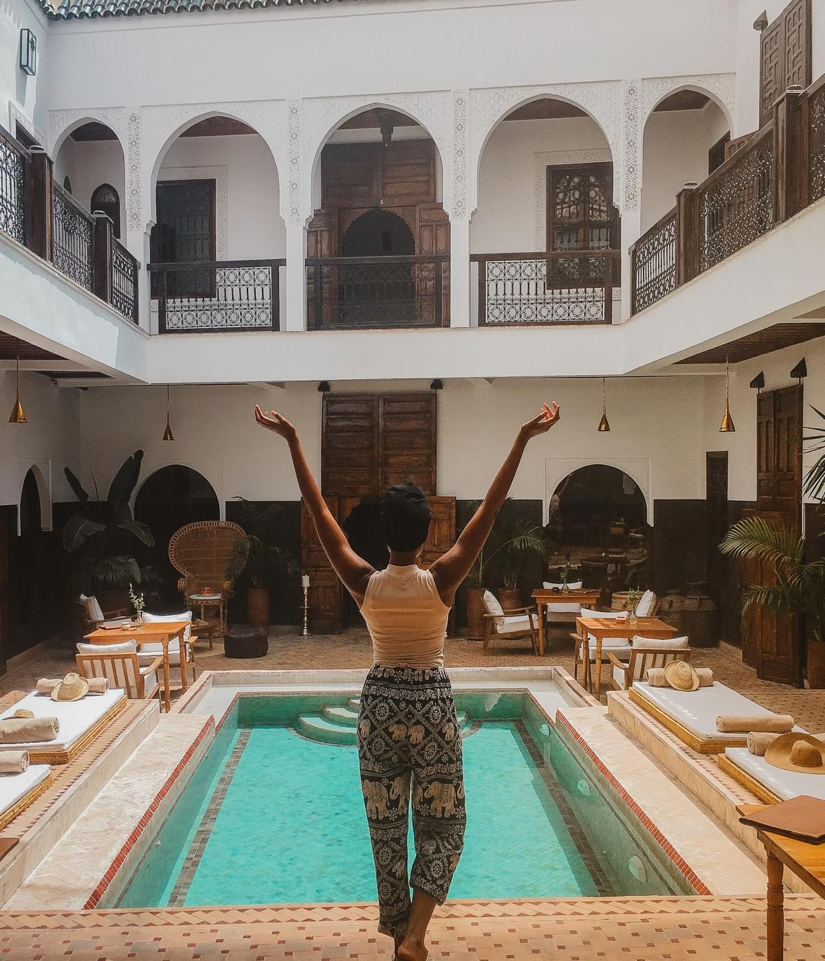 Black woman with raised arms in front of a pool in Riad Kasbah courtyard with open doors and mats and tables Black woman with raised arms in front of a pool in Riad Kasbah courtyard with open doors and mats and tables