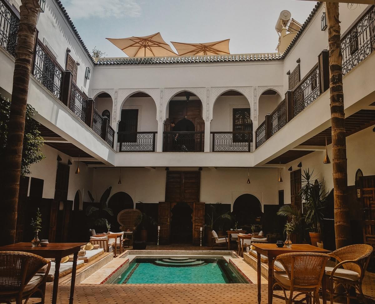 The courtyard of a traditional road with tables, chairs, mats, and plants surrounding a pool and open umbrellas on the roof The courtyard of a traditional road with tables, chairs, mats, and plants surrounding a pool and open umbrellas on the roof