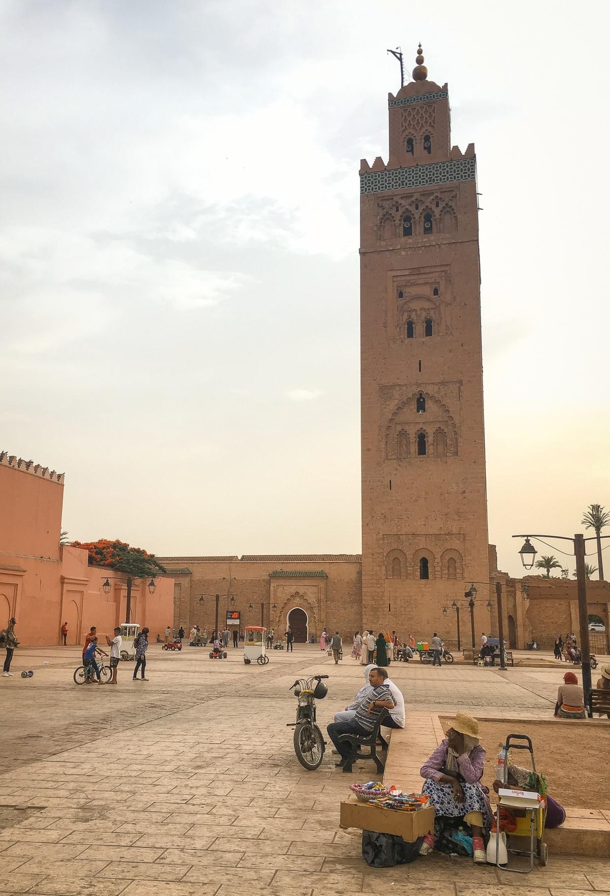 Woman selling goods in open medina with men sitting on a bench and bikes near the minerat of El Mouassine Mosque Woman selling goods in open medina with men sitting on a bench and bikes near the minerat of El Mouassine Mosque
