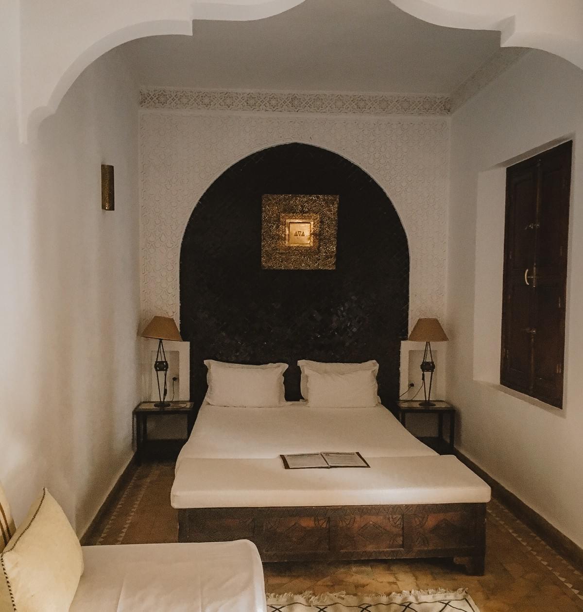 A room with white couch and pillows, a double bed with side tables and lamps on either side. with Moroccan detailing, crown molding, and gold center photo and closed window A room with white couch and pillows, a double bed with side tables and lamps on either side. with Moroccan detailing, crown molding, and gold center photo and closed window