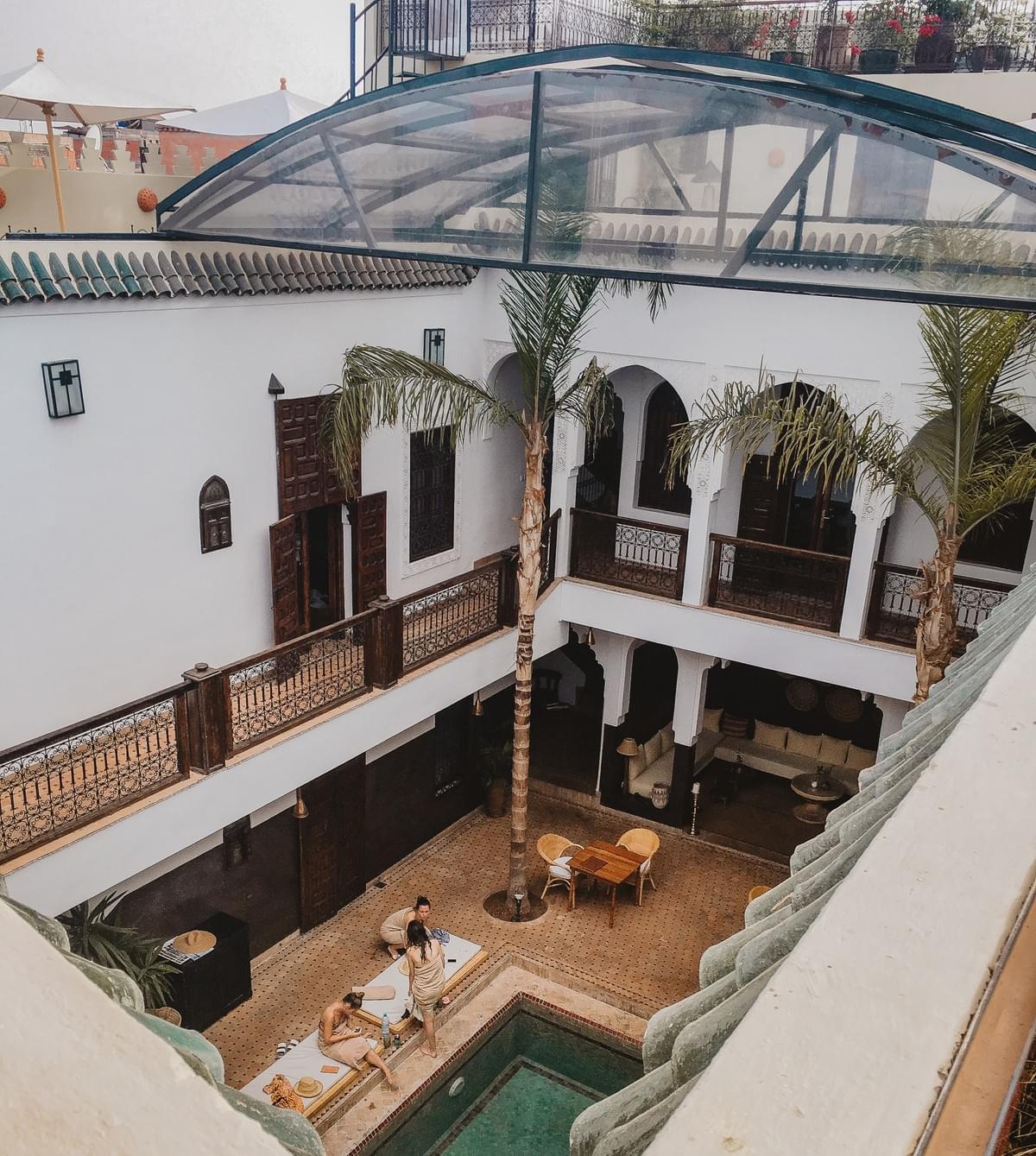 Three girls sitting by a courtyard pool with a table and two palm trees and brown rails and clear awning at Riad Kasbah Three girls sitting by a courtyard pool with a table and two palm trees and brown rails and clear awning at Riad Kasbah