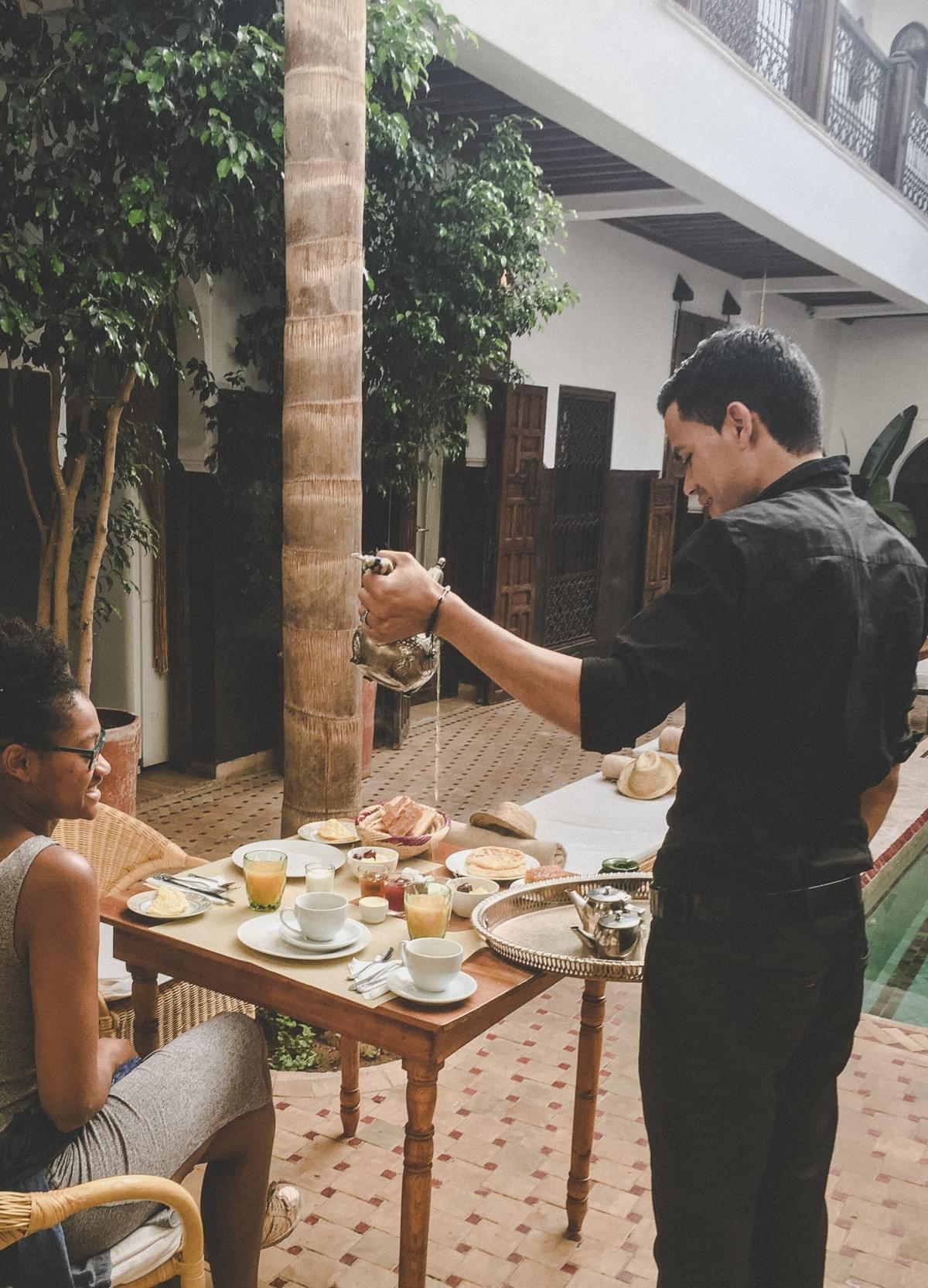 back girl with natural hair and grey dress sitting at outdoor table with breakfast watching a male waiter pouring nana tea out of antique moroccan teapot back girl with natural hair and grey dress sitting at outdoor table with breakfast watching a male waiter pouring nana tea out of antique moroccan teapot