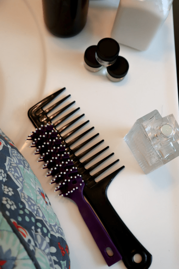 a black comb, purple brush, perfume bottle, vera bradley toiletry bag, glass jars and lotion on a bathroom counter a black comb, purple brush, perfume bottle, vera bradley toiletry bag, glass jars and lotion on a bathroom counter