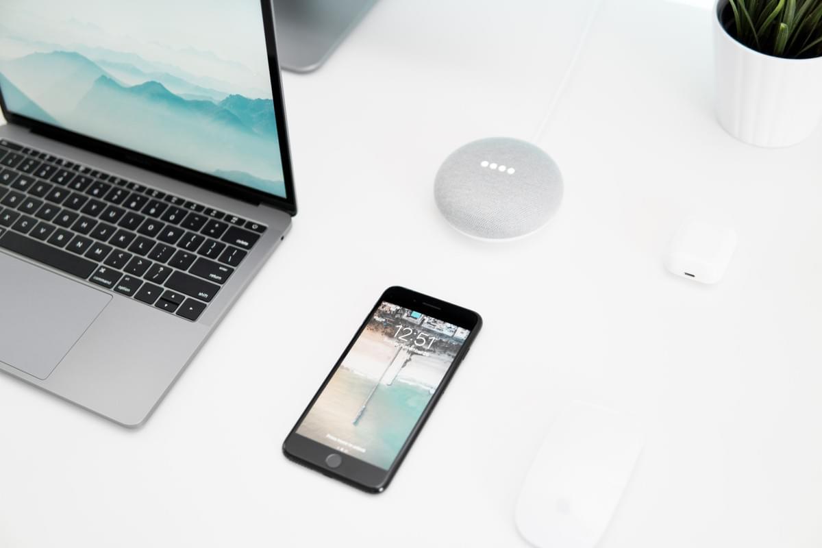 un computer macbook, un iphone ed un dispositivo google home su una scrivania bianca un computer macbook, un iphone ed un dispositivo google home su una scrivania bianca