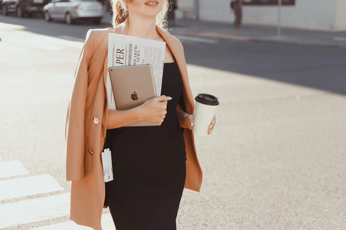 Professional woman holding laptop, newspaper, and coffee as she crosses the street