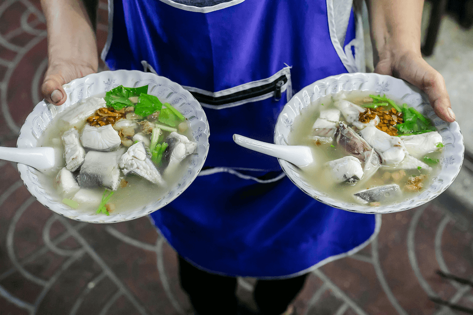 Close-up of a vendor holding two bowls of clear fish soup (likely khao tom pla or similar Southeast Asian dish) containing white fish fillets, herbs, and crispy fried garlic.