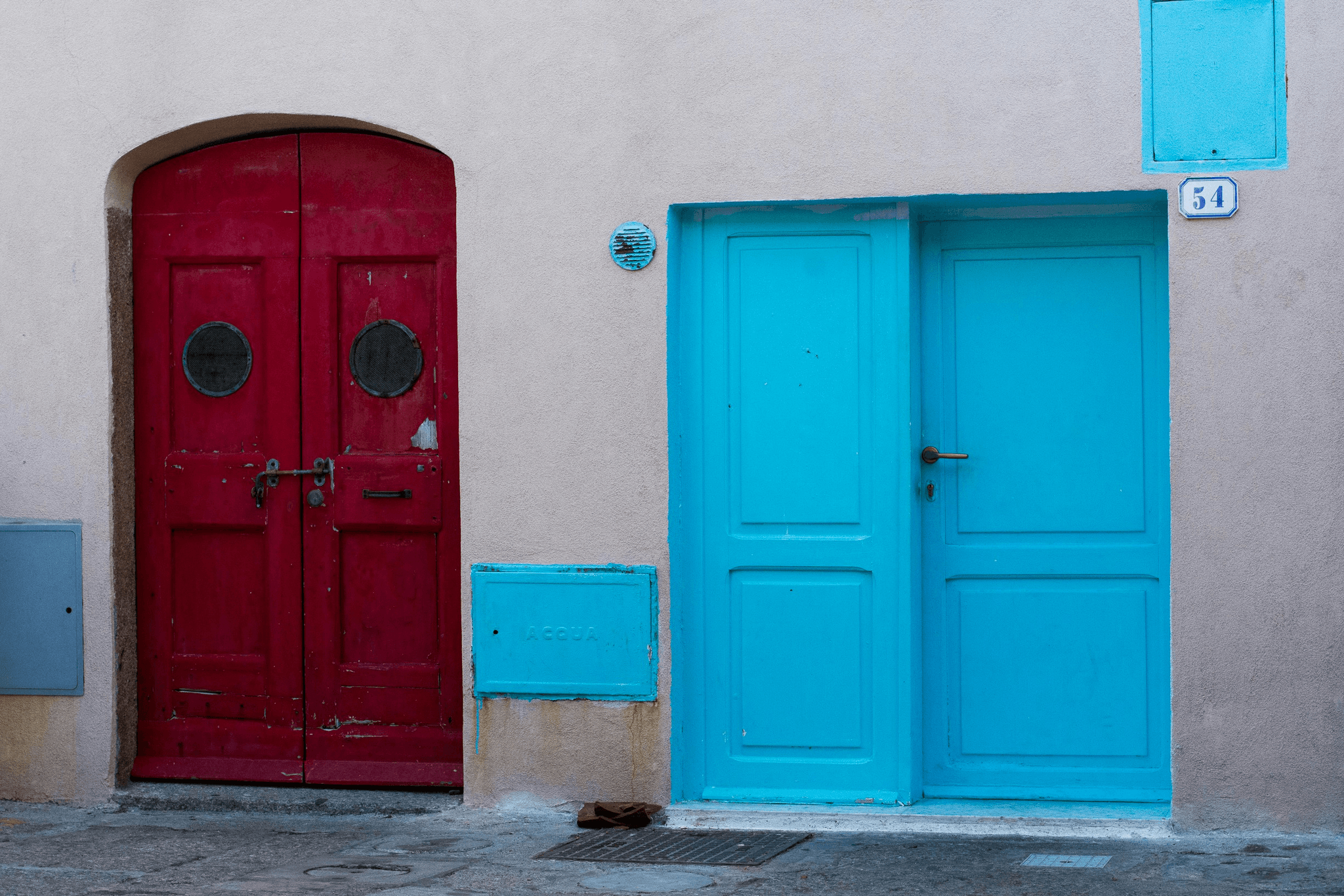 Two contrasting doors on a beige wall: a deep red door with circular windows and a central metal latch beside a bright turquoise door with a brass handle and matching utility panels labelled “ACQUA.” The bold red‑and‑blue colour contrast creates a striking, graphic architectural scene.