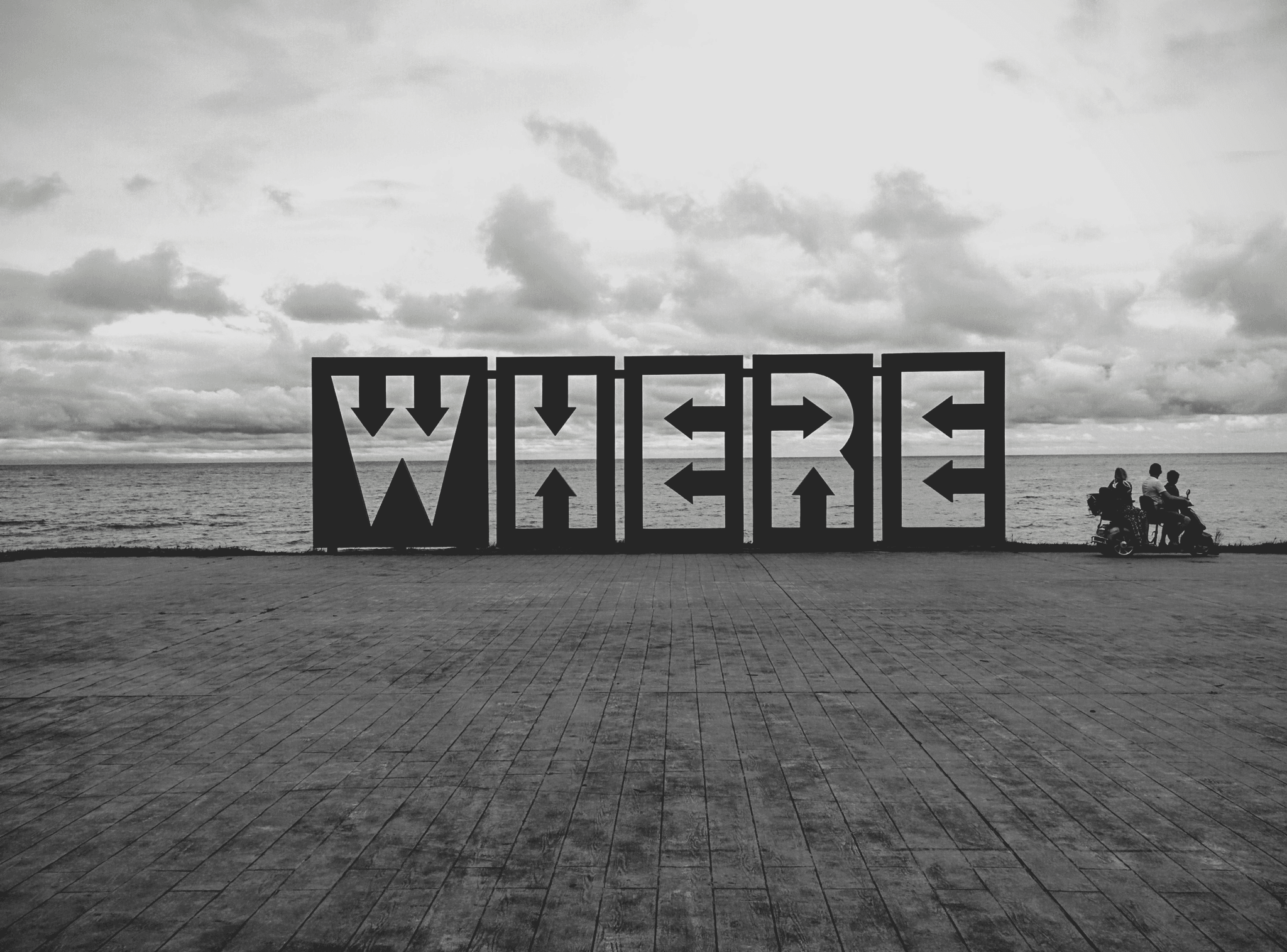 A black-and-white photograph of a beachside art installation composed of six large blocks arranged in a straight line, each displaying a bold capital letter to spell the word “WHERE.” The blocks face the viewer directly, creating a striking visual prompt. In the background, a body of water stretches beneath a cloudy sky, suggesting a coastal location. To the right of the installation, two people are positioned near bicycles, adding a human element to the contemplative scene. The image evokes themes of inquiry, place, and existential reflection, with the word “WHERE” serving as both a literal and symbolic question.