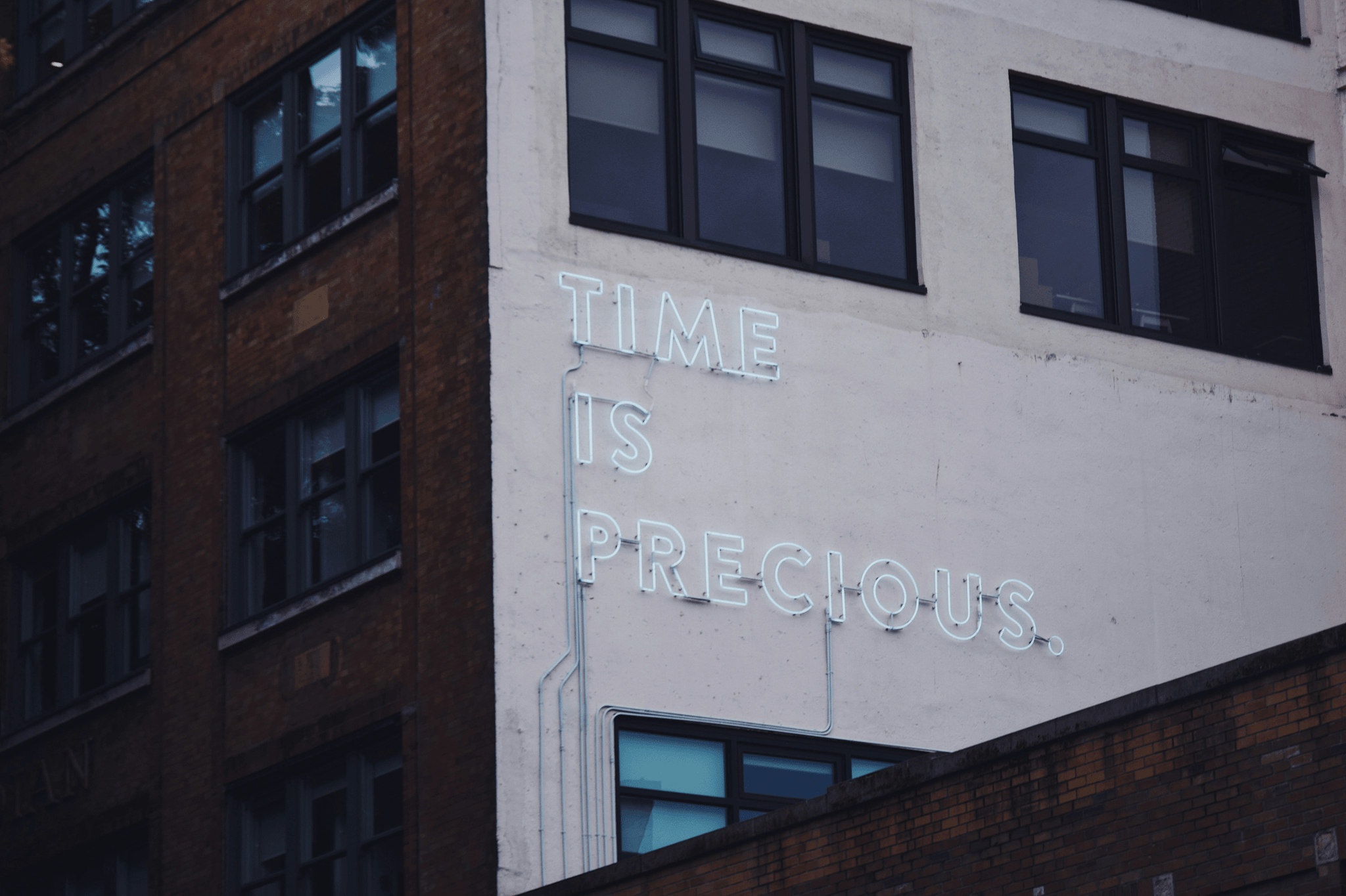 Corner view of a multi-story building featuring a bold neon sign that reads “TIME IS PRECIOUS” in glowing white letters. The sign is mounted on a white-painted section of the facade, contrasting with the adjacent brown brickwork and rows of windows. The building’s architectural mix of textures—brick, paint, glass—adds urban character, while the illuminated message stands out as a philosophical prompt or public reminder. The composition captures a moment of stillness and reflection in the city, suggesting themes of urgency, mindfulness, and the value of presence amid everyday routines.