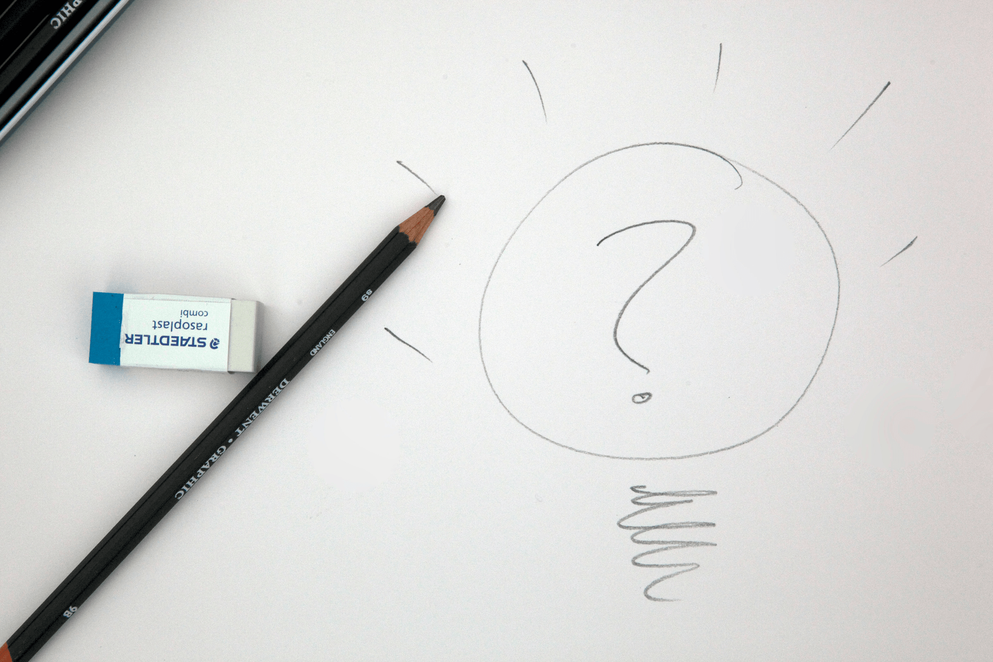 A pencil and a white eraser rest on a sheet of paper featuring a hand-drawn sketch of a light bulb with a question mark inside it. The bulb is outlined with simple lines, radiating short strokes at the top to suggest illumination. Inside, a scribbled filament anchors the question mark, symbolizing curiosity or the spark of an idea. The composition is minimal yet evocative, blending tactile tools of creation with a visual metaphor for inquiry, brainstorming, or creative problem-solving. The image invites reflection on the process of thinking, learning, and generating insight.