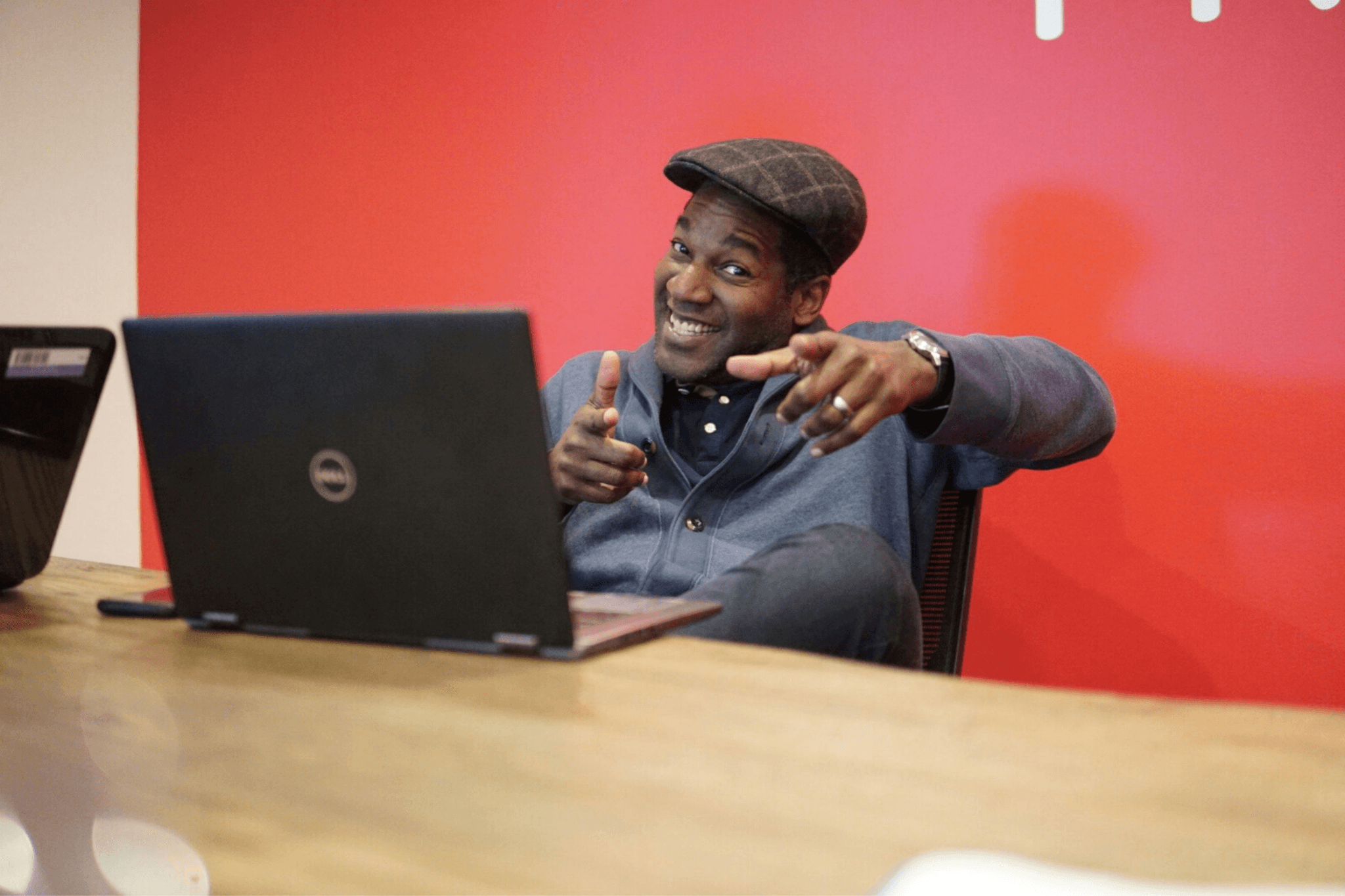 A person sits at a table in front of an open Dell laptop, smiling and pointing both index fingers forward in a lively, engaging gesture. They wear a dark flat cap and a blue cardigan, and the background is a solid red wall that adds warmth and energy to the scene. The setting feels informal and welcoming, possibly a casual workspace or meeting area. The person’s expressive posture and cheerful demeanor convey enthusiasm, encouragement, and approachability—suggesting a moment of connection, affirmation, or light-hearted motivation.