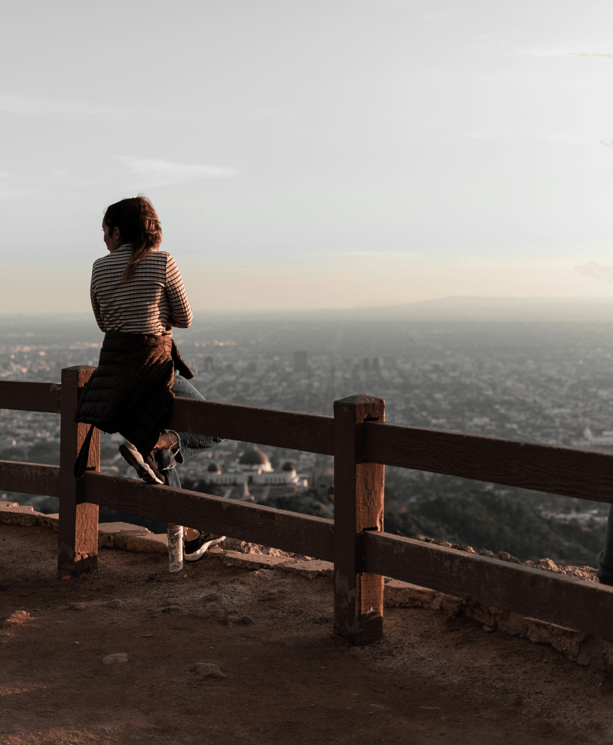 A casually dressed person sits alone on a wooden fence at a scenic overlook, gazing out over a sprawling cityscape below. The individual wears a striped shirt, jeans, and sneakers, with a dark jacket tied around their waist. A water bottle rests on the ground nearby, suggesting a pause during a walk or hike. The viewpoint is elevated—likely a hill or mountain—offering a panoramic view of densely packed urban buildings stretching toward the horizon. The sky above is hazy, casting a soft, muted light over the scene. The composition conveys a quiet moment of reflection and solitude, with the contrast between the natural foreground and the vast, structured city below evoking themes of contemplation, escape, and connection between nature and urban life