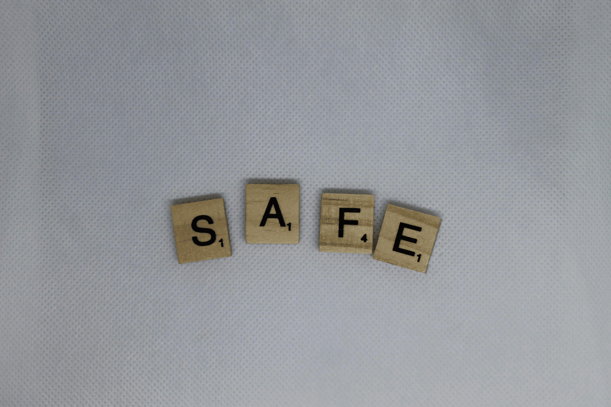 The word “SAFE” is gently spelled out using four wooden Scrabble tiles, each letter carrying a quiet weight: S (1), A (1), F (4), E (1). The tiles sit on a soft white surface, evoking calm and clarity. Their warm tones and simple arrangement suggest comfort, stability, and the small but meaningful steps we take toward feeling secure. A visual whisper of reassurance — safety built letter by letter