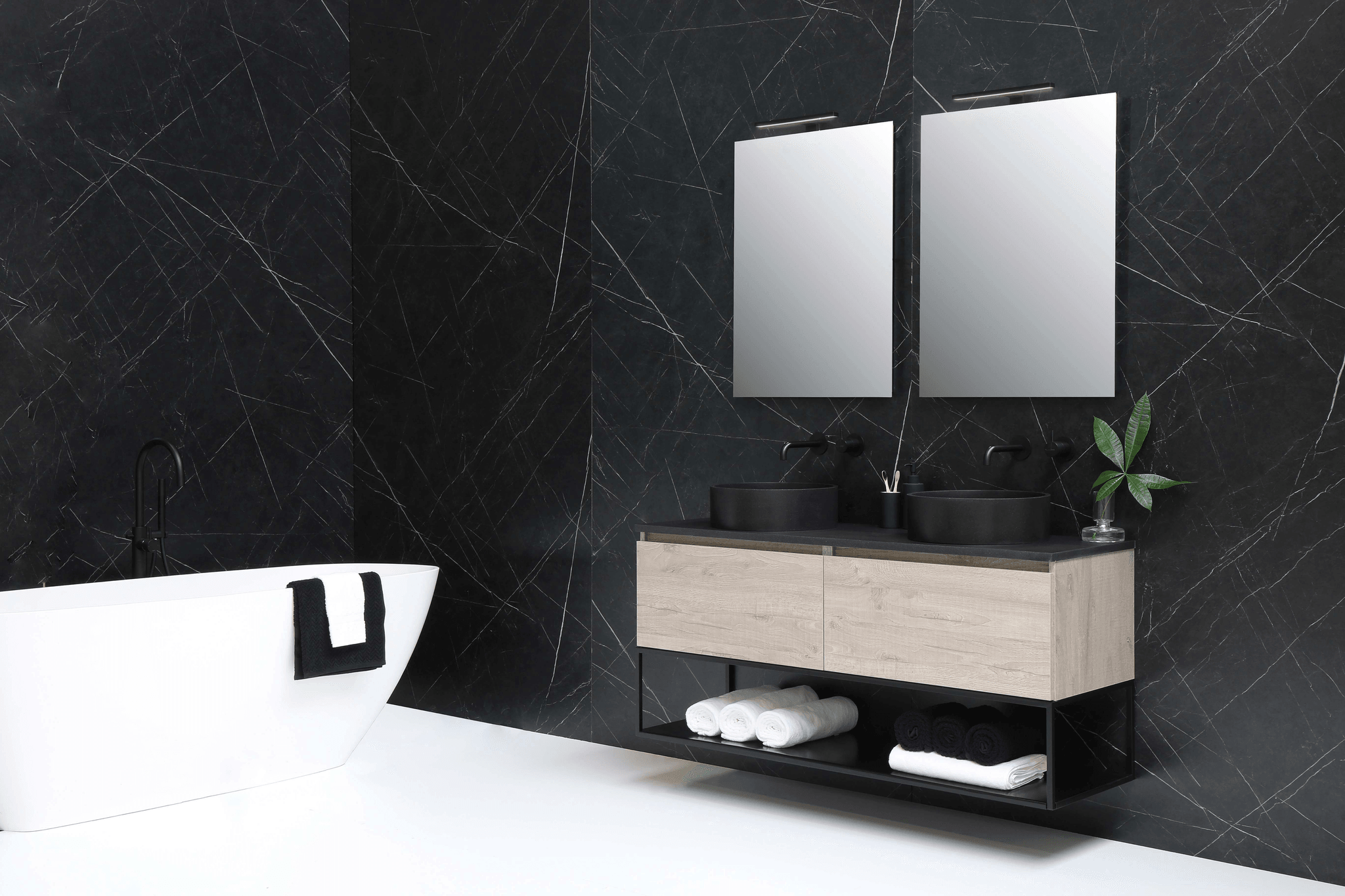 Bathroom with a white bathtub with a black and white towel. A wooden style vanity with towels underneath, double basin and double square mirrors placed on black and gold tiles.