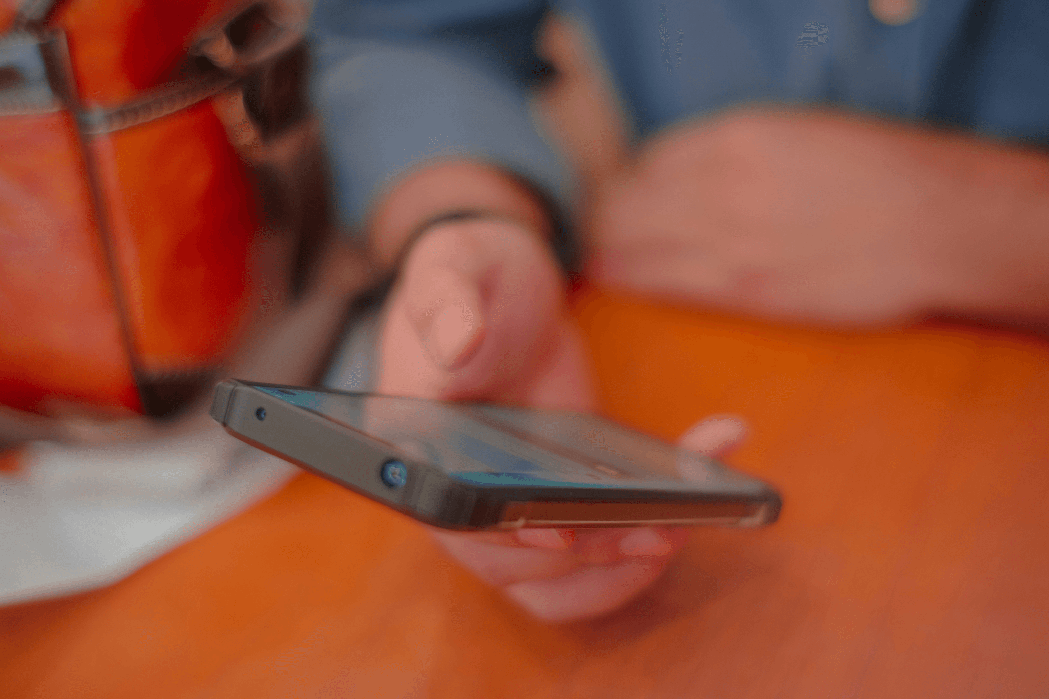 “A person holding a smartphone above a wooden table, with part of a blue sleeve and a brown leather bag visible in the background.