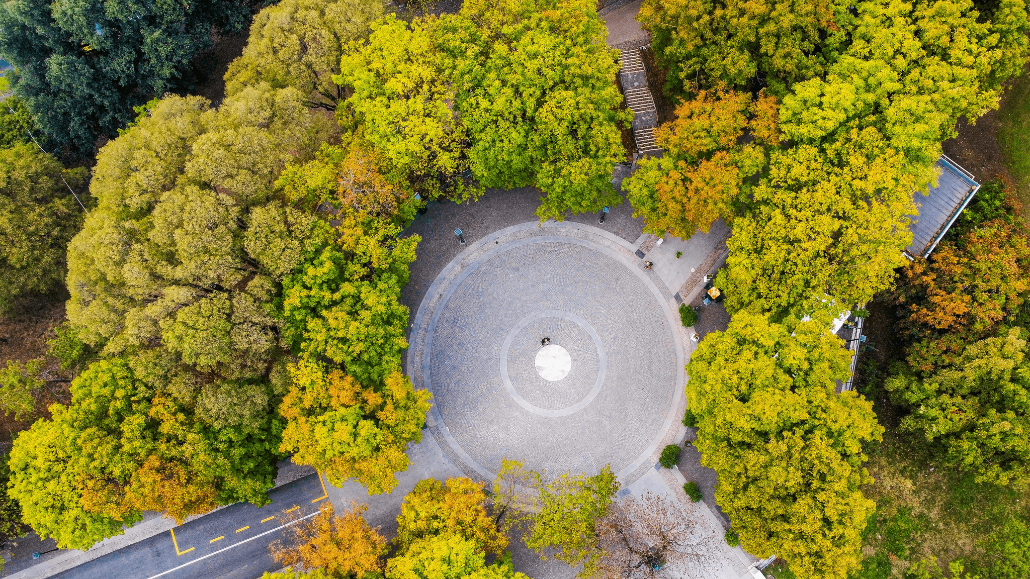    An aerial view of a circular plaza surrounded by dense, multicolored foliage, showcasing a seasonal transition with trees in shades of green, yellow, and orange—likely indicating early autumn. The plaza features a central white structure, possibly a sculpture or fountain, encircled by concentric paving patterns that radiate outward. Pathways and a road with yellow markings are visible on the left side, while a building partially obscured by trees appears in the top right corner. The symmetrical layout and vibrant natural palette create a visually striking composition, blending urban design with environmental beauty. The image evokes themes of balance, seasonal change, and thoughtful landscape architecture.